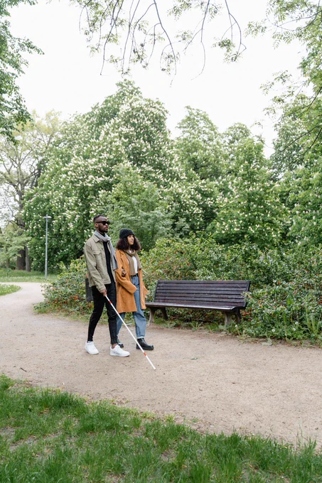 We see a photograph of a visually impaired male accompanied by his female carer/partner as they enjoy a walk in a peaceful and beautiful park.