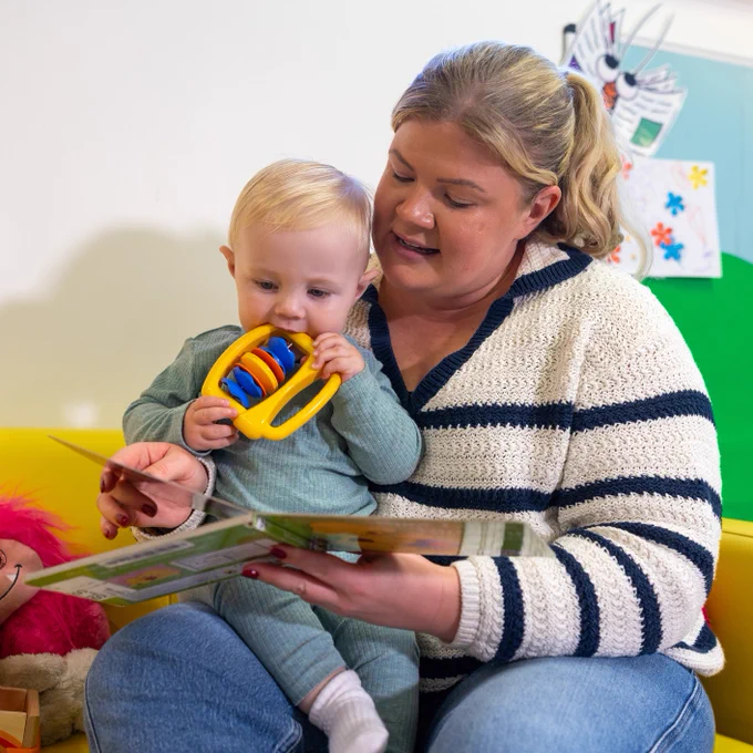 A woman reads a book to a baby on her lap, who is chewing on a toy. They are sitting on a yellow sofa.