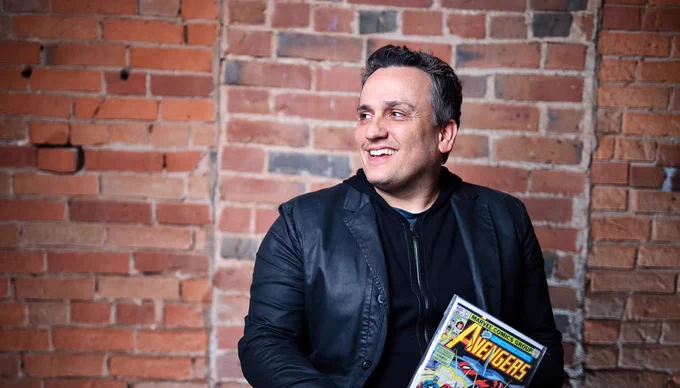 Joe Russo wearing a black jacket and dark shirt sitting against a brick wall, holding a vintage Avengers comic book.