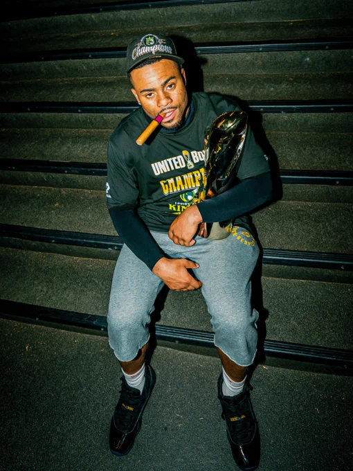A person sits on stairs wearing a championship cap and shirt, holding a trophy and a cigar. The mood is celebratory and triumphant.