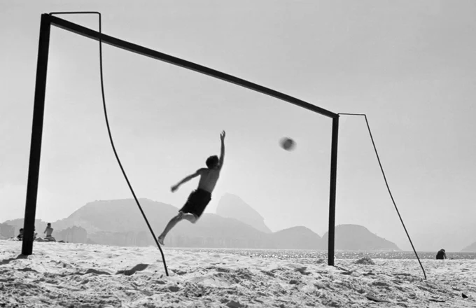 Fotografia in bianco e nero che mostra un ragazzo sospeso a mezz'aria sulla spiaggia di Copacabana a Rio de Janeiro mentre si tuffa per parare un pallone da calcio, incorniciato da una porta da calcio artigianale.