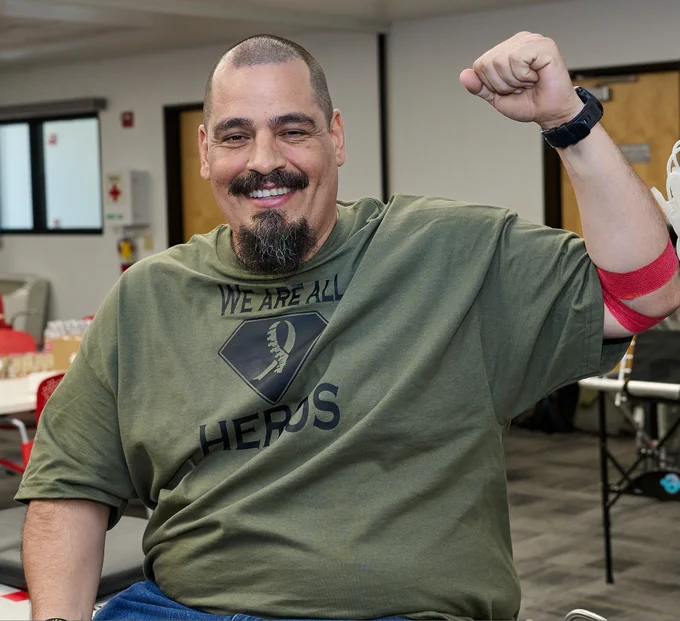 A person smiling and flexing an arm while seated in a room. They are wearing a green shirt with the text "We Are All Heroes" and a bandage is visible on their arm. The room has tables and chairs.