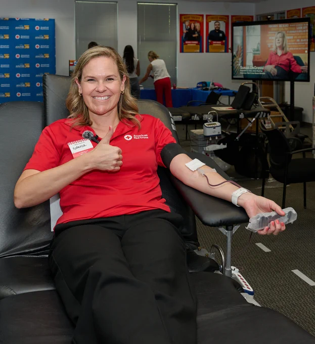 An individual wearing a red polo shirt is sitting on a donation chair, giving a thumbs-up while donating blood. A Red Cross badge is visible on the shirt. In the background, there are Red Cross posters and a screen displaying an image of another person speaking.