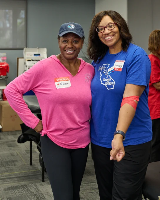 Two people smiling and standing together in a room with chairs and boxes. One person is wearing a pink shirt and cap with a volunteer name tag, and the other is wearing a blue shirt with a California design, and has a bandage on an arm.