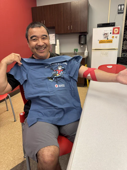 The person is seated indoors, smiling, and holding up a blue T-shirt with an American Red Cross design. There's a red bandage on their arm. The background features a red and gray wall, wooden cabinets, and a white refrigerator with various magnets.