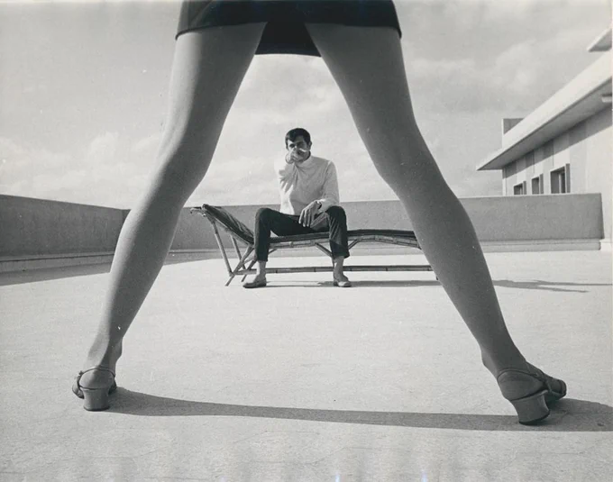 Fotografia in bianco e nero all'attore George Lazenby nei panni di James Bond. L'inquadratura dal basso è incorniciata in primo piano dalle gambe divaricate di una donna che indossa una minigonna e scarpe con il tacco, creando una prospettiva geometrica a triangolo. Sullo sfondo, al centro della composizione, George Lazenby è seduto su un lettino prendisole su una terrazza sul tetto, mentre fuma una sigaretta e guarda dritto verso l'obiettivo.