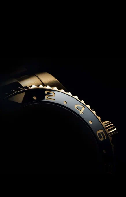 Macro, high-contrast photograph of a gold Rolex GMT-Master II bezel emerging from deep shadows