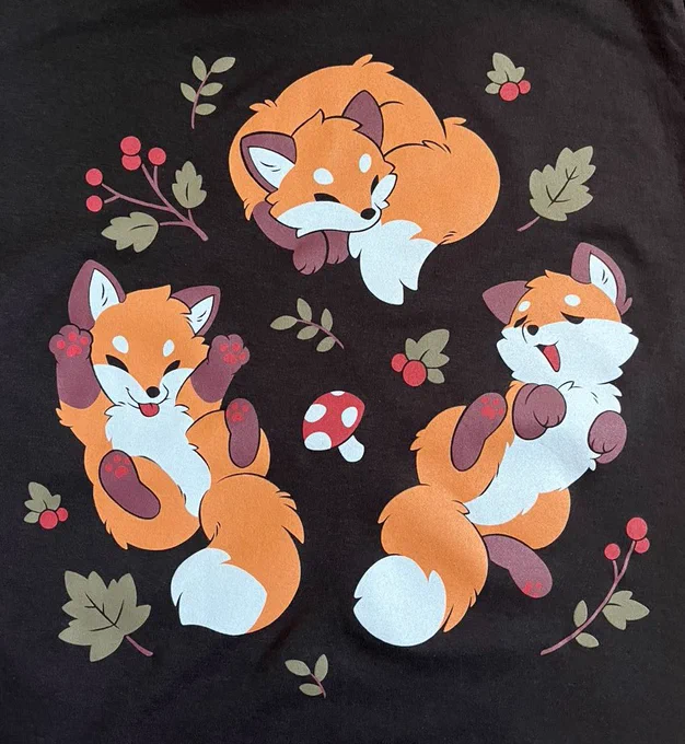 A shirt with a fluffy fox design. They are in various playful poses, and they are surrounded by plants and mushrooms.