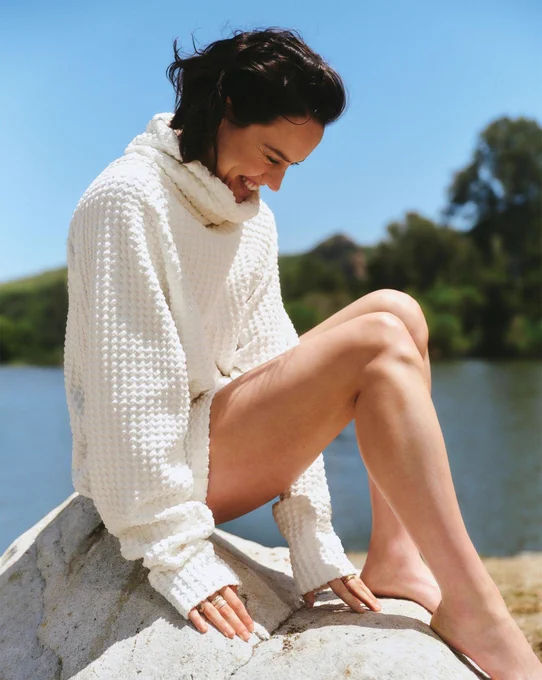 L'attrice Daisy Ridley siede di profilo su una roccia chiara all'aperto, sorridendo spontaneamente con lo sguardo rivolto verso il basso. Indossa un morbido maglione oversize bianco sporco con una spessa trama a nido d'ape e un ampio collo a dolcevita. Le gambe sono nude e i piedi scalzi, poggiati sulla pietra. Sullo sfondo si notano uno specchio d'acqua calmo e alberi sfocati sotto un cielo blu limpido.