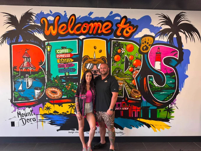 A vibrant wall mural in Mount Dora featuring colorful elements like palm trees, a lighthouse, and fruits. Two people stand smiling in front of the mural, creating a cheerful and lively atmosphere.