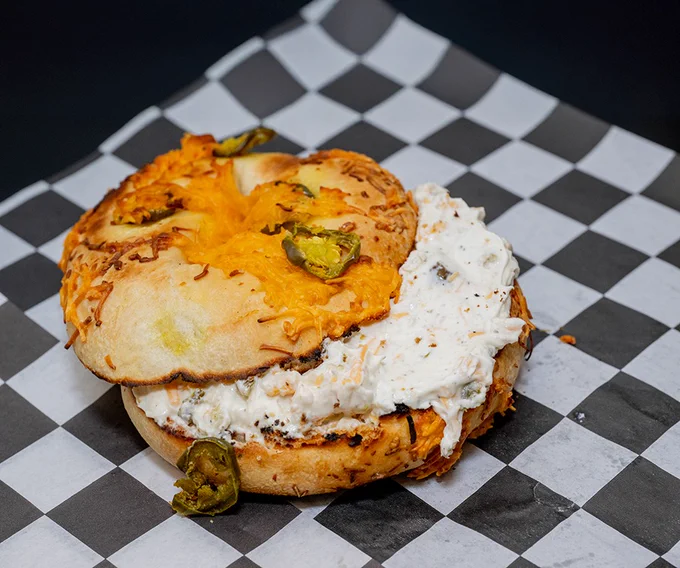 A close-up of a jalapeño and cheese bagel filled with cream cheese on a black and white checkered paper. The bagel appears freshly baked with melted cheese on top.