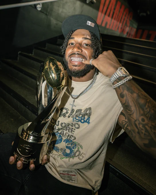 Athlete smiling and showcasing a championship trophy while pointing to his mouth, highlighting his grill. The setting is casual and celebratory.