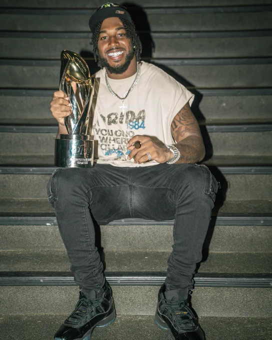 A man with a bright smile sits on stairs holding a shiny trophy. His casual attire and accessories add a relaxed yet cheerful mood.