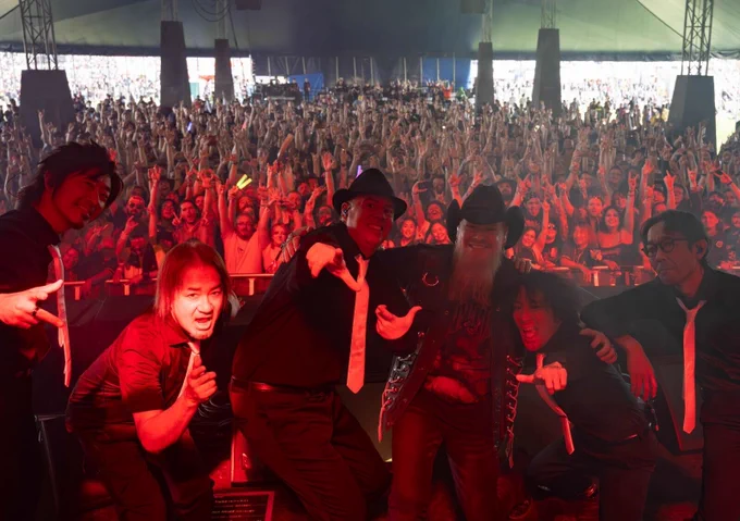 A photograph of THE PRIMALS and Jason Charles Miller posing on stage with their backs facing the crowd. Many members of the audience are cheering while doing the rock on gesture.