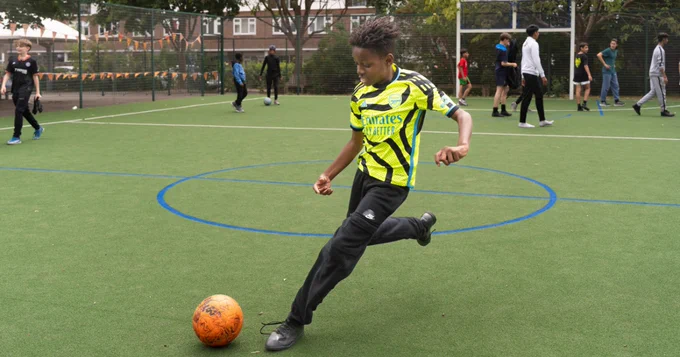 A child kicking an orange football on an outdoor sports pitch with green artificial turf. Several other children and teenagers are visible in the background near a wire fence and a basketball hoop.