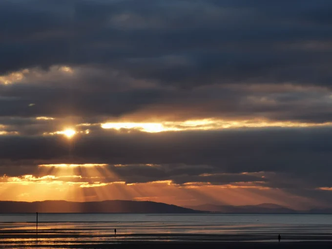 Sun setting behind clouds over Snowdonia (Eryri) looking across the Irish Sea from Crosby beach, near Hall Road train station on Merseyrail's Southport line.

The scene is composed of a wide shot of the beach and sea. The horizon line is clearly defined, separating the sky from the water. The sun, positioned near the horizon, is radiating golden light, casting rays that pierce through the clouds. In the background, a low, distant horizon line reveals the silhouette of land the mountain range. The clouds are various shades of grey, dark grey, and hints of orange and yellow, creating a dramatic contrast with the golden sunlight. The foreground shows the flat, sandy beach with statues from Antony Gormley's 'Another Place' installation. The middle ground reveals the calm water of the sea, extending to the shoreline.