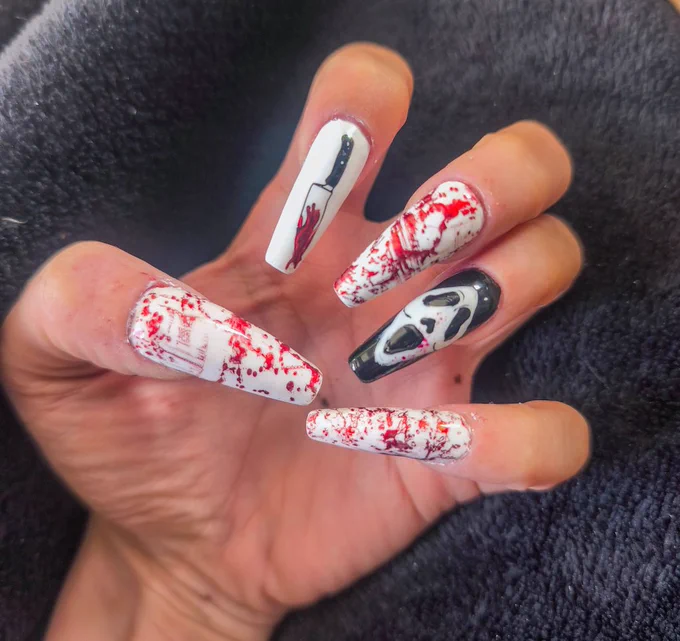 > A close-up photograph of a hand displaying long coffin-shaped acrylic nails with a horror-themed design.

The manicure uses a predominantly white, black, and red color palette. Several nails feature red splatter effects across a white base, creating the appearance of paint or blood-like staining. Fine line details and distressed textures add depth to the design.

One accent nail depicts a black-handled knife with a red-stained blade against a white background. Another features a stylized white ghost mask with elongated black eye and mouth openings set against a black background.

Additional nails incorporate red splatter patterns and faint graphic details layered over the white base, giving the overall set a worn, cinematic horror aesthetic.

The glossy finish reflects light across the surface of the nails, emphasizing the contrast between the bright white background, the deep black elements, and the vivid red accents.

The hand is positioned against a dark fabric background, drawing