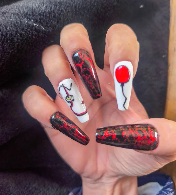 A close-up photograph of a hand displaying long coffin-shaped acrylic nails with a horror-inspired design.
Three of the nails feature a black base with vivid red marbling and crack-like patterns, creating a textured, distressed appearance. The glossy finish enhances the contrast between the black and red elements.
Two accent nails use a white base. One nail depicts part of a stylized face with a yellow eye, black eyeliner, and red facial markings. The other features a bright red balloon outlined in black, with a thin black string extending toward the tip of the nail.
The color palette is dominated by black, red, and white, creating a strong visual contrast. The combination of the painted eye, facial details, and red balloon motif gives the manicure a cinematic horror aesthetic.
The image focuses entirely on the nail artwork, with the hand positioned against a dark fabric background that helps emphasize the intricate details and bold colors of the design.