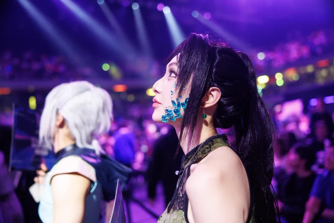 Two people in cosplay stand in a brightly lit arena, watching the VCT Masters London broadcast. One person in the foreground has black hair and blue crystal-like face decorations, while another with white hair stands beside them.