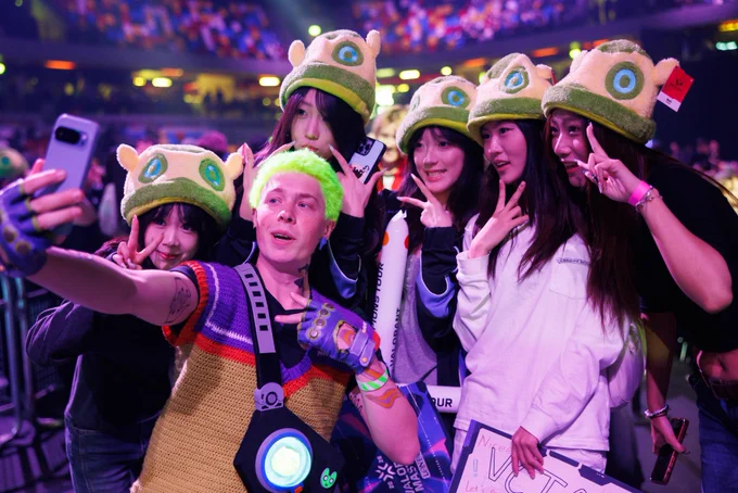 A group of fans wearing matching Mosh hats gather for a selfie with a Gekko cosplayer in a crowded arena, smiling and posing for the camera.