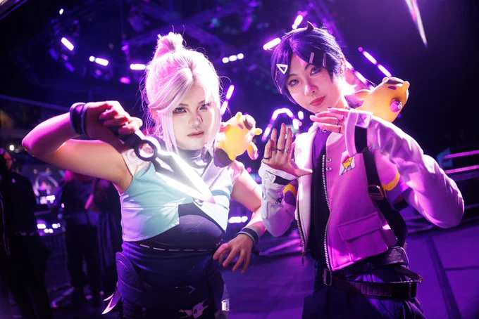 Two people in Jett and Clove cosplays pose under purple stage lights at the arena, with one holding a glowing prop melee while both face the camera.