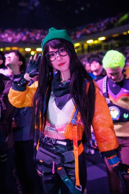 A person in a Killjoy cosplay poses and waves at the camera in a crowded arena, wearing a green beanie, large round glasses, and a yellow jacket.