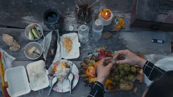 A still from the film How To Feed A Dictator. Food is being prepared on a rough, aged wooden surface. In one corner, light brown hands are at work. There are scattered pieces of old crockery, small spoons, shrimp, dragon fruit, grapes, sticky rice etc.