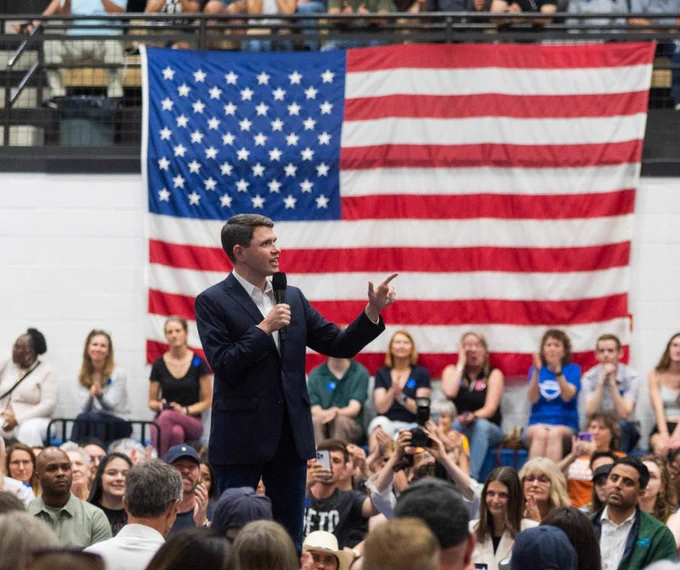 Photo of James Talarico speaking at a rally with a large American flag hanging in the background.