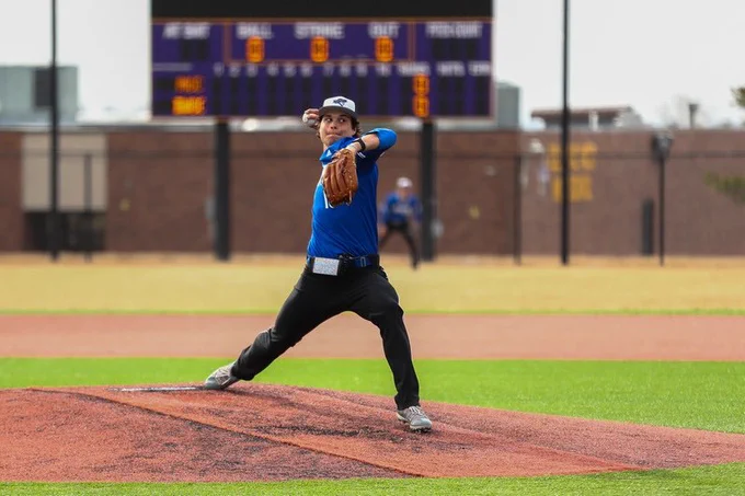 A right handed baseball pitcher throws a pitch from the mound with a scoreboard in the background