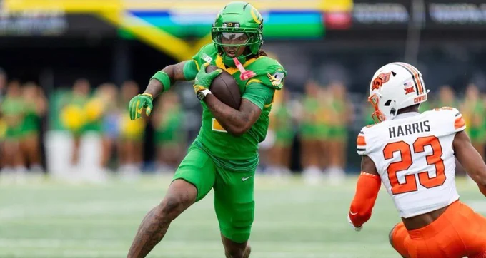 Football player in green uniform runs with the ball as a defender in white and orange closes in from the right.