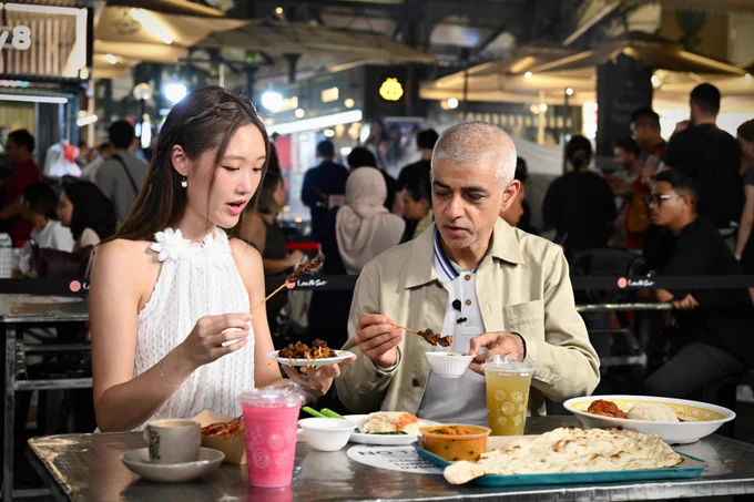 Mayor of London Sadiq Khan and guide sit at a busy outdoor food market table at night, each holding skewers of grilled food above small plates. The table is covered with various dishes including flatbread, curry, rice, and colourful drinks in plastic cups. Bright stall lights illuminate a crowded background filled with diners, umbrellas, and signage.