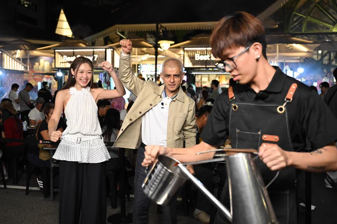 In a lively night market setting, Mayor of London Sadiq Khan and guide stand near a food stall while a worker in the foreground pours a drink between two metal containers. 