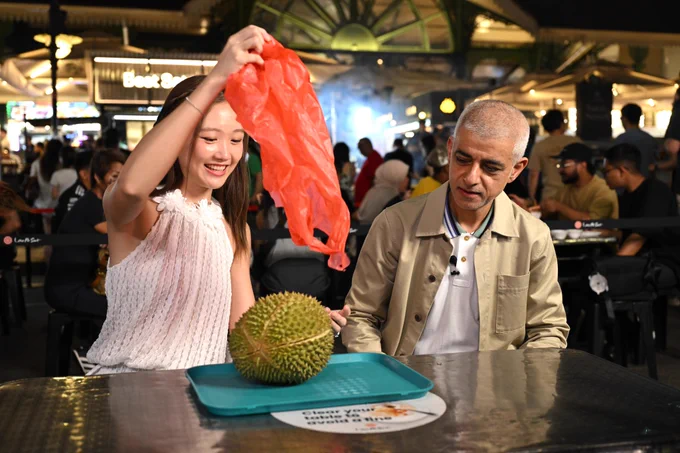 At a bustling night market table, a guide lifts a red plastic bag away from a large spiky durian fruit resting on a tray, while Mayor of London Sadiq Khan looks toward the fruit. 