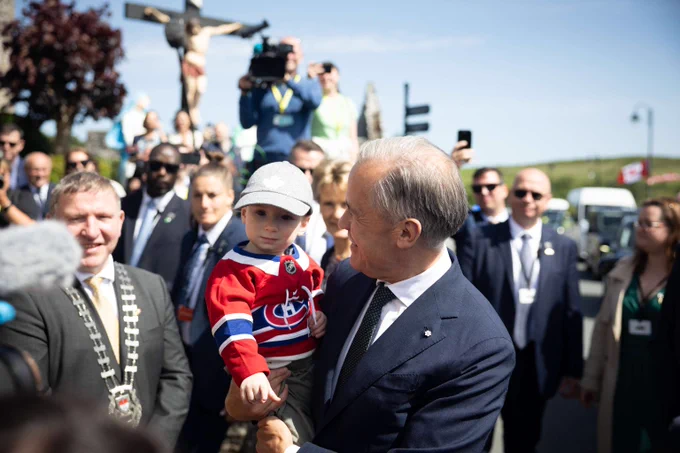 Le premier ministre Mark Carney rencontre des membres de la communauté d’Aughagower dans le comté de Mayo, en Irlande. Il tient dans ses bras un enfant portant un chandail des Canadiens de Montréal.