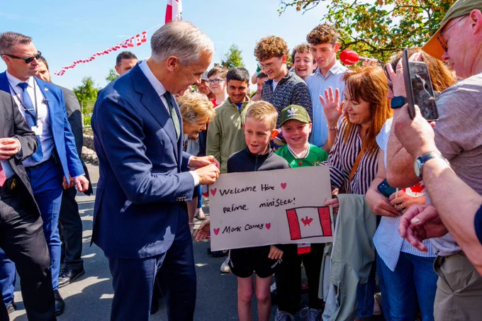 Le premier ministre Mark Carney rencontre des membres de la communauté d’Aughagower dans le comté de Mayo, en Irlande. Il regarde en souriant l’affiche tenue par un enfant, sur laquelle on peut lire : « Bienvenue à la maison, Monsieur le Premier ministre, Mark Carney ».