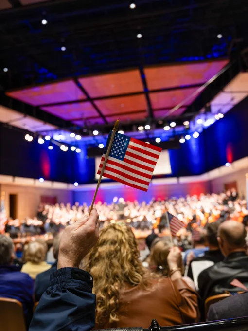 A person in the audience holds up an American flag inside a concert hall. The stage is brightly lit and features a choir and orchestra performing. The ceiling has wooden acoustic panels, and the audience is seated in rows.