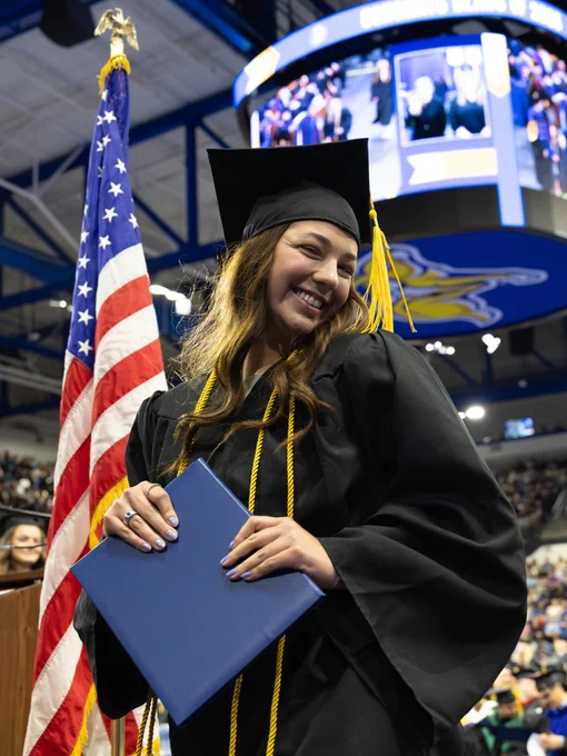 A graduate in a cap and gown holds a diploma cover while standing beside an American flag. A large screen in the background displays a ceremony scene. The venue is filled with people attending the event.