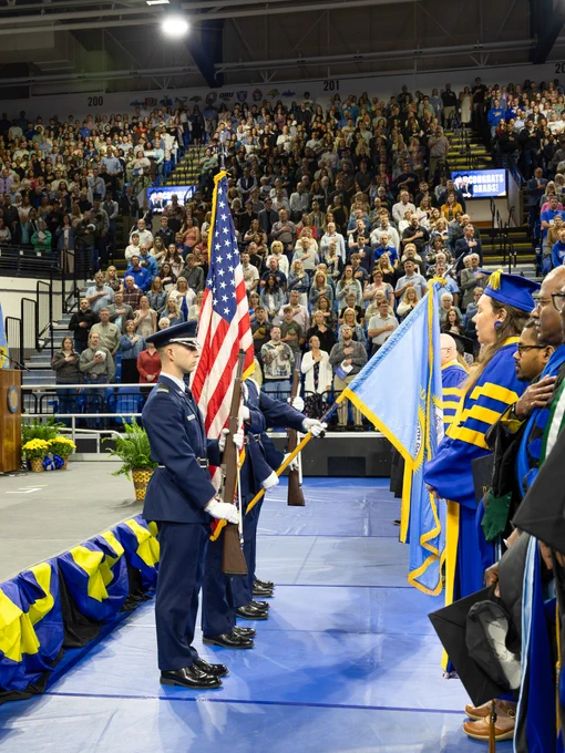 A graduation ceremony in a large indoor arena. A group of uniformed individuals stands holding flags, including the American flag, near the stage. Graduates in blue and yellow academic regalia face the flag bearers. The audience, filling the stands, watches the proceedings.