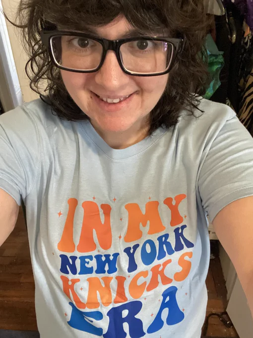 Me, a white woman with black-framed glasses. I’m wearing a light blue shirt that reads, “In my New York Knicks Era.”