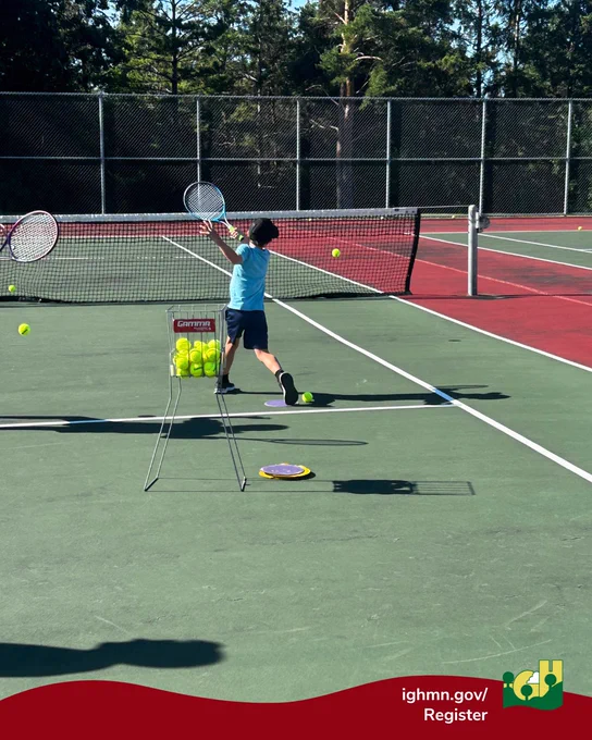 Young boy practicing tennis serves on an outdoor court with a basket of tennis balls nearby.