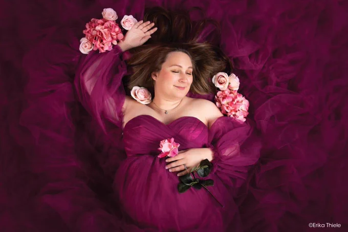 A person peacefully lying on a flowing deep magenta fabric, surrounded by pink roses. Copyright Erika Thiele.