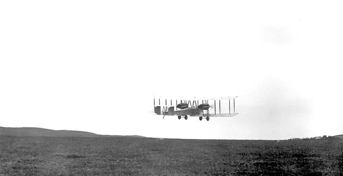 A vintage biplane is flying low over an open field, silhouetted against a bright sky.