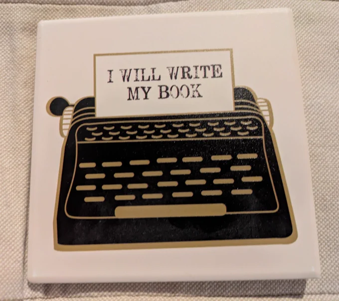 Black typewriter drawing with gold keys on square ceramic white coaster. Paper emerging from the typewriter says: I WILL WRITE MY BOOK