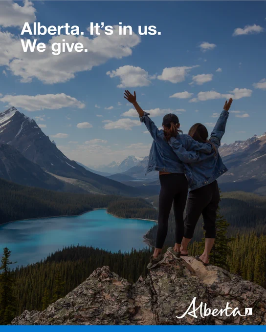 Two people stand on mountain top and raise their arms to the sky as they look at the view. Text reads: “Alberta.It’s in us. We give.” Alberta logo is in bottom right corner. 