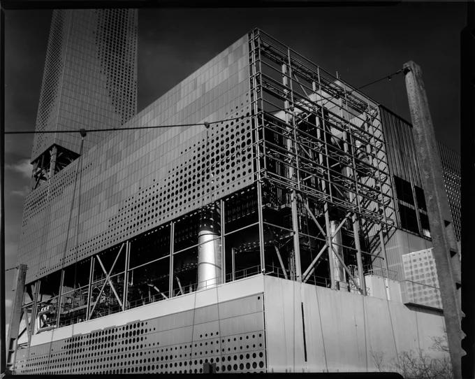 black and white picture of a large industrial building with a seemingly metallic panelling in front of a complex assortment of tubes and conduits. the panelling has a few different shades of grey in the picture, and is pierced with a lot of round holes in a wavy pattern. on the right side, we can see the metal structure that supports the upper levels of the building and through it the inner workings of its function, and on the left is a tall tower covered in the same panelling