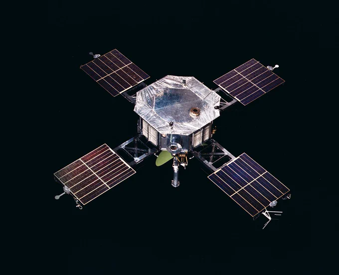 A spacecraft with a central octagonal body and four extended solar panels. The surface appears metallic, with various instruments attached. The background is the blackness of space.