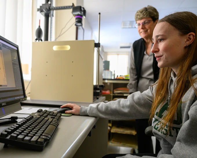 Two people are in a lab setting. One is sitting at a desk using a computer with a scanning device nearby, while the other stands behind, observing and smiling. There are shelves and equipment in the background.