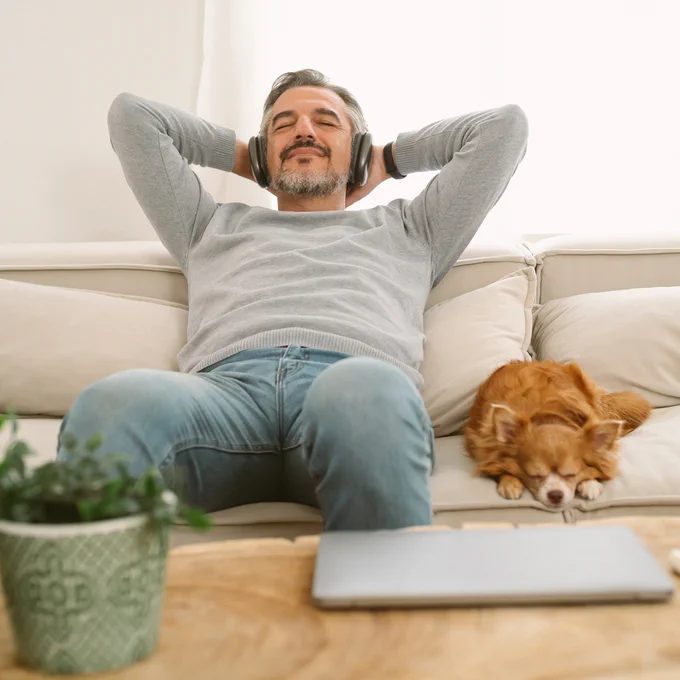 A man sitting on a couch wearing headphones with a small dog beside him.