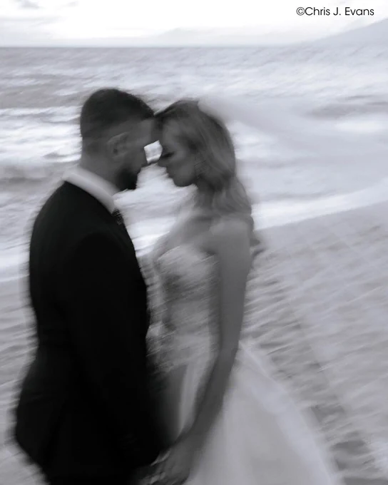 This is a black and white, blurred photograph of two people standing close together on a beach. The ocean and waves are visible in the background. Copyright Chris J. Evans.