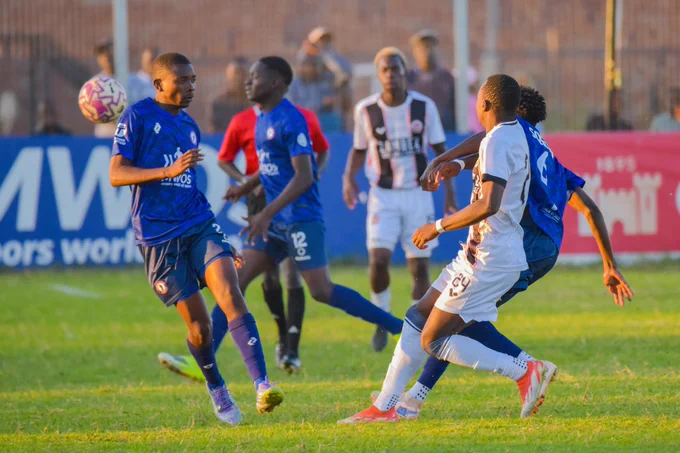 Players from two teams compete for the ball during a soccer match, wearing blue and white uniforms on a green field in the sunlight.